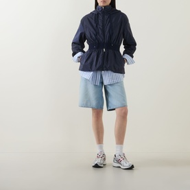 Model wearing Moncler Liriope Logo Raincoat in navy with drawstring waist, styled with blue striped shirt, denim shorts and white trainers.