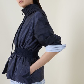 Model wearing Moncler Liriope Logo Raincoat in navy, showing elasticated waist and side pockets, styled with blue striped trousers.