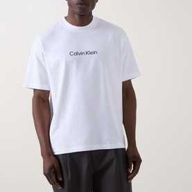 Model wearing Calvin Klein Standard Logo T-Shirt in Bright White, crew neck with short sleeves and chest logo print, styled with dark trousers.