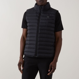Model wearing Calvin Klein Logo Padded Gilet in black, sleeveless with standing collar and zip fastening, styled with black T-shirt and trousers.