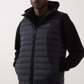 Model wearing Calvin Klein Logo Padded Gilet in black, sleeveless with standing collar and zip fastening, styled over a black long-sleeve top.