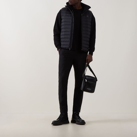 Model wearing Calvin Klein Logo Padded Gilet in black over black hoodie and trousers, carrying a black shoulder bag with black shoes.