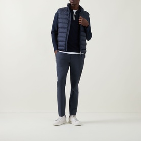 Model wearing Calvin Klein Tech Stretch Slim-Leg Trousers in Dark Sapphire, styled with a navy quilted gilet, navy knit and white trainers.