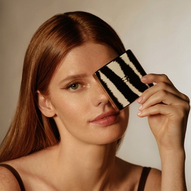 Model with Sisley Phyto-Eye Palette in Tender Toffee, zebra-print compact held up to eye, black strap top visible