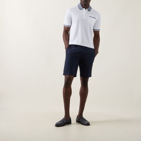Model wearing Bugatti Contrast Collar Polo Shirt in white with navy tipped collar and cuffs, styled with navy shorts and dark loafers.