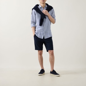 Model wearing Bugatti Lightweight Drawstring Shorts in navy, styled with a blue striped shirt, navy jumper over shoulders, and navy canvas trainers.