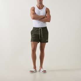 Model wearing Moncler green swim shorts with drawstring waist and small logo detail, styled with a white vest and white slides.