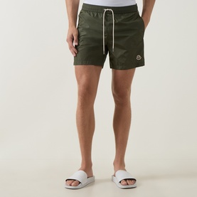 Model wearing Moncler green swim shorts with drawstring waist and logo badge, styled with white slider sandals.