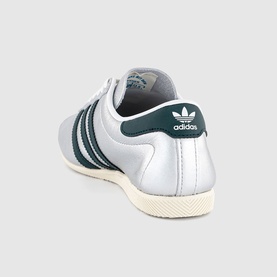 Adidas trainer in silver metallic with dark green three-stripe detailing, tonal heel tab, and cream sole, rear angle view.