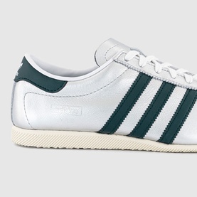 Adidas trainer in silver metallic with dark teal three stripes, lace-up fastening, and cream rubber cupsole.