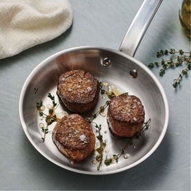 Tramontina silver stainless steel frying pan with three seared beef medallions, fresh thyme, and sea salt flakes