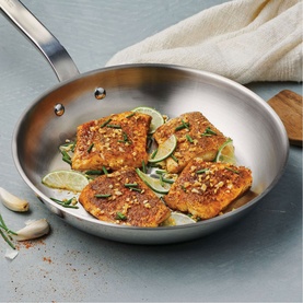 Tramontina silver stainless steel frying pan with seared salmon fillets, lime slices, and garlic, styled on a grey surface