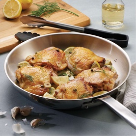 Tramontina silver stainless steel frying pan with cooked chicken thighs, styled with chopping board, tongs, and garlic on grey surface