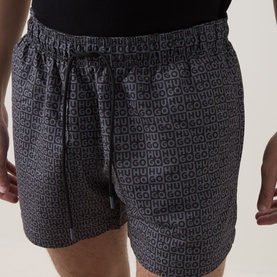 Model wearing Hugo black swim shorts with all-over monogram logo print, elasticated waistband and drawstring tie.