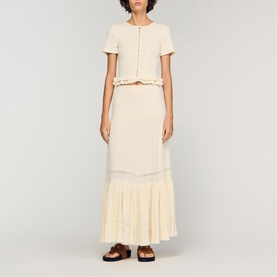 Model wearing Sandro Ruffle Knit Cardigan in Clear, cropped with pearl buttons and ruffled sleeves, styled with matching maxi skirt and brown sandals.
