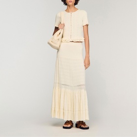 Model wearing Sandro Ruffle Knit Cardigan in clear, styled with matching maxi skirt, carrying a cream bag, wearing brown sandals.