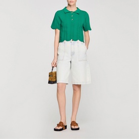Model wearing Sandro Scallop Trim Knitted Top in emerald green with scallop hem, styled with white shorts and brown sandals, carrying small box bag.