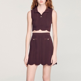 Model wearing Sandro Wave Cropped Knit Top in brown with buttoned polo collar, sleeveless, styled with matching brown scallop-hem mini skirt.