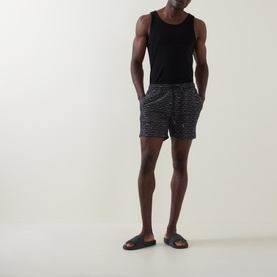 Model wearing Hugo Boss black swim shorts with all-over print, styled with a black vest top and brown slider sandals.