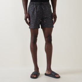 Model wearing Hugo Boss black all-over logo print swim shorts, styled with navy slider sandals.