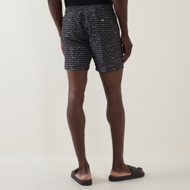 Model wearing Hugo Boss black swim shorts with all-over text print, shown from behind, styled with black slider sandals.