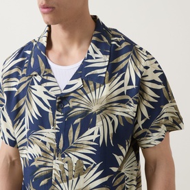 Model wearing Hugo Boss dark beige and navy tropical leaf print short-sleeve shirt with camp collar, styled over a white crew-neck top.