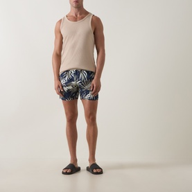 Model wearing Hugo Boss dark beige palm-print swim shorts with a beige vest top and dark brown slider sandals.