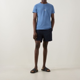 Model wearing Hugo Boss light blue T-shirt with small chest logo, styled with navy swim shorts and brown slider sandals.