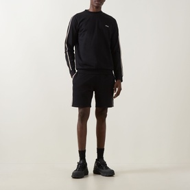 Model wearing Hugo Boss black cotton sweatshirt with white shoulder stripe detailing, styled with matching black shorts and chunky trainers.