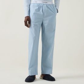 Model wearing Calvin Klein Logo Gingham Pyjama Bottoms in ballad blue plaid, elasticated logo waistband, straight-leg fit, styled with navy slippers.