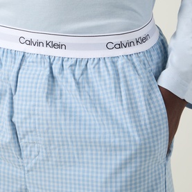 Model wearing Calvin Klein Logo Gingham Pyjama Bottoms in Ballad Blue check print with white elasticated logo waistband, paired with white tee.