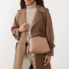Model carrying Gianni Chiarini Alifa Crossbody Bag in clay hammered leather, silver-tone hardware, slim shoulder strap. Styled with camel trench coat.