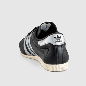 Adidas trainer in core black and silver metallic leather, three-stripe detailing, white heel tab, cream rubber sole, rear angle view.