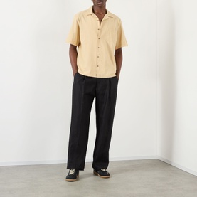 Model wearing A.P.C. Jude Pocket Shirt in beige with bowling collar and short sleeves, styled with black trousers and dark trainers.