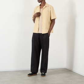 Model wearing A.P.C. Jude Pocket Shirt in beige with bowling collar, short sleeves, styled with black wide-leg trousers and dark loafers.