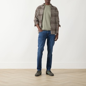 Model wearing Seven Jeans Army crew neck T-shirt in sage green, styled with plaid overshirt, blue slim jeans and grey suede shoes.