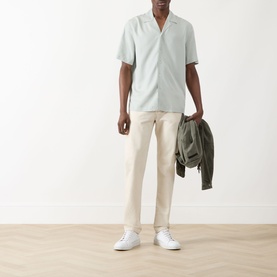 Model wearing Seven Jeans Dusty Blue lyocell reg shirt, styled with cream trousers and white trainers, carrying olive jacket