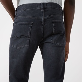 Model wearing Seven Jeans Slimmy Slim Leg Jeans in Breakwater, showing rear five-pocket detail, logo badge, and faded denim finish.