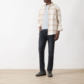 Model wearing Seven Jeans Slimmy Slim Leg Jeans in Breakwater, styled with a cream plaid overshirt and light blue tee, grey suede shoes.