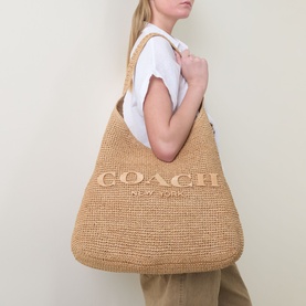 Model carrying Coach Brooklyn Weave Shoulder Bag 39 in natural paper straw, hobo silhouette with tonal logo, paired with white tee and khaki trousers
