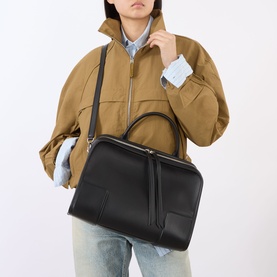 Model carrying Loewe Amazonia 180 Mini Top Handle Bag in black calfskin with silver-tone hardware and crossbody strap, styled with camel jacket
