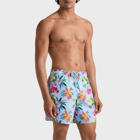 Model wearing Vilebrequin swim shorts in Fleur de Lin, featuring a vibrant tropical floral print with palm trees on a light blue ground.