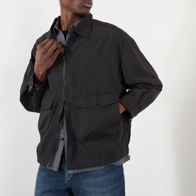 Model wearing Edwin Chaos Water-Resistant Jacket in black, zip fastening, patch pockets with flap, styled with blue jeans and checked shirt.