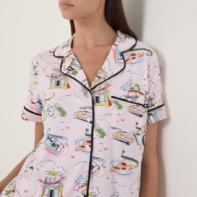Model wearing Kate Spade City Stroll pyjama top in pink with colourful city-scene print and contrast navy piping, short-sleeved shirt collar style.