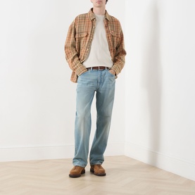 Model wearing Nudie Jeans Tuff Tony Straight Leg Jeans in Drift Wood, styled with a plaid overshirt, cream tee, and brown leather shoes.