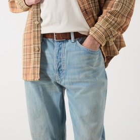Model wearing Nudie Jeans Tuff Tony Straight Leg Jeans in Drift Wood, loose fit with faded whiskering detail, styled with brown belt and plaid shirt.