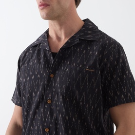 Model wearing Nudie Jeans Reg Shirt in R22 Multi, black ikat-print short-sleeve camp collar shirt with brown buttons, close-up detail view.