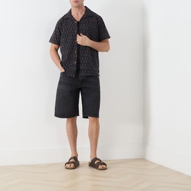 Model wearing Nudie Jeans Reg Shirt in R22 Multi, short sleeve camp collar with allover print, styled with black denim shorts and brown sandals.