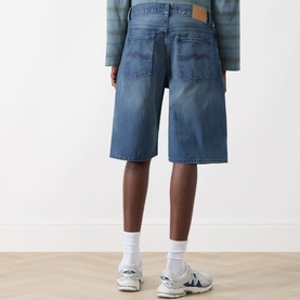 Model wearing Nudie Jeans Zeb High-Rise Wide Leg Denim Shorts in Blue Painter, rear view, showing logo badge, faded details and wide-cut legs.