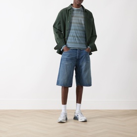 Model wearing Nudie Jeans Zeb High-Rise Wide Leg Denim Shorts in Blue Painter, styled with striped tee, green overshirt and white trainers.